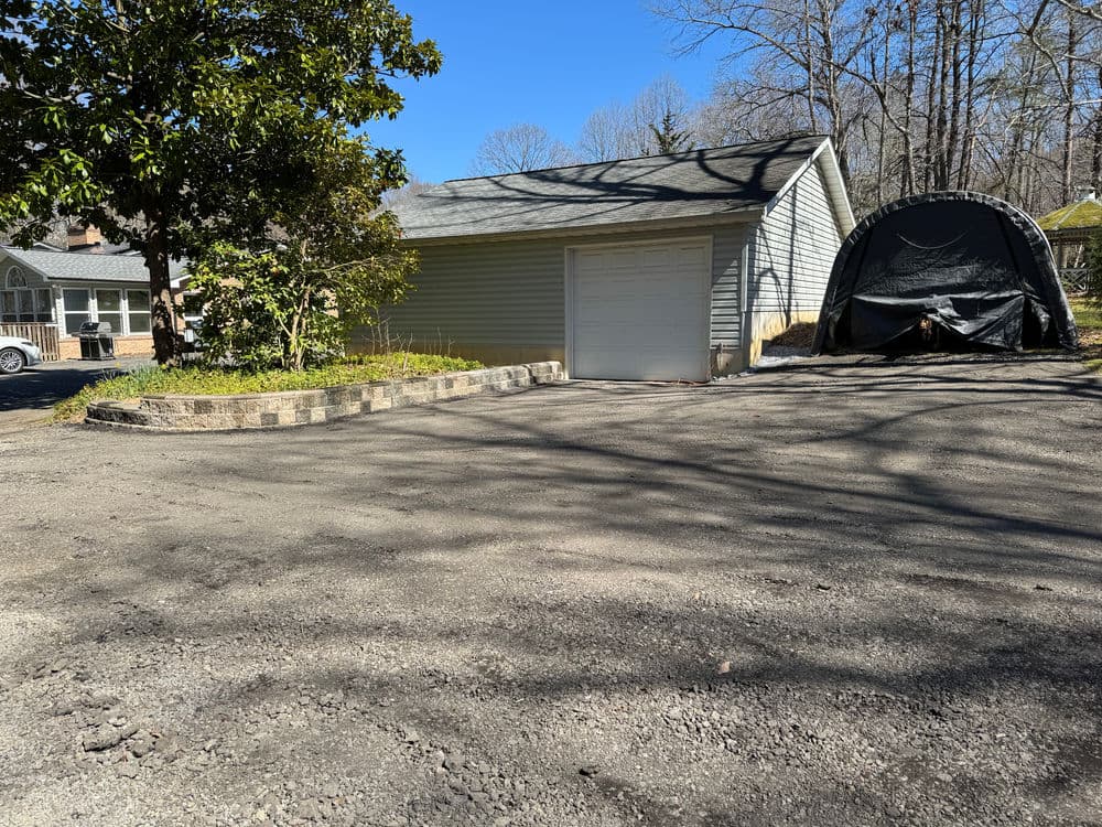 A gravel driveway leading to a gray garage with a portable shelter nearby in a wooded area.
