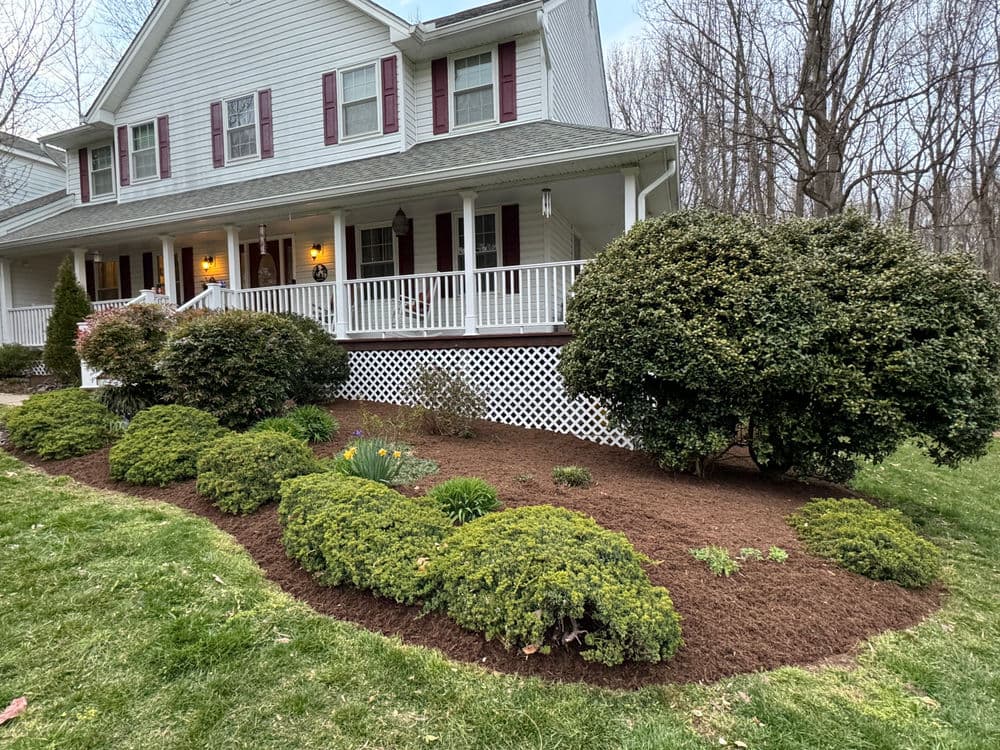 Front yard landscaping with shrubs, mulch, and a white porch of a two-story house.