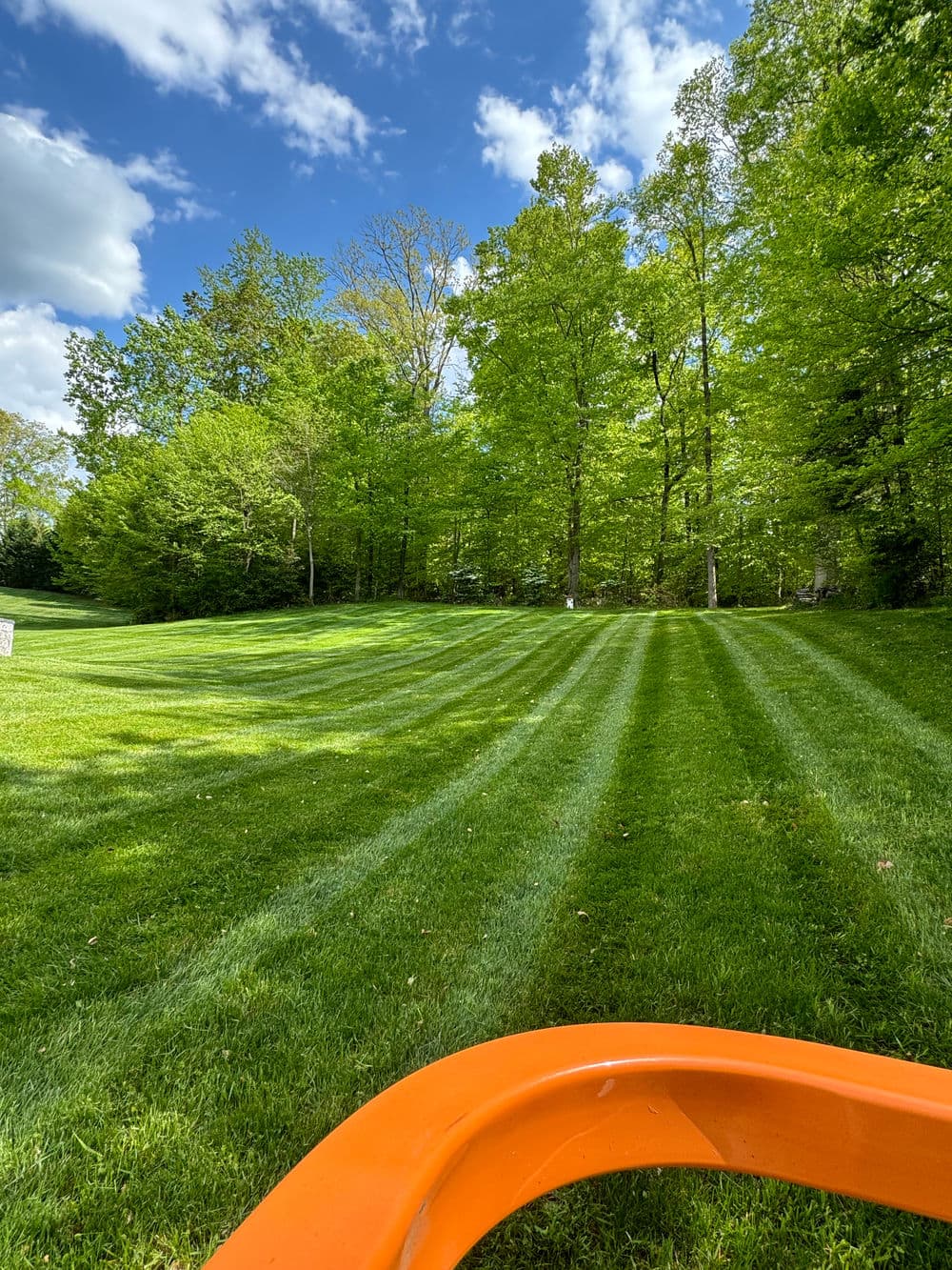 Lush green lawn with freshly mowed stripes under a bright blue sky and scattered clouds.